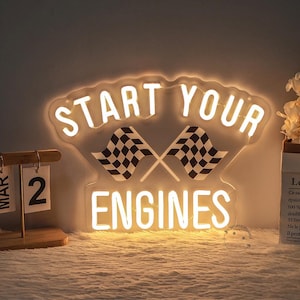 May include: A neon sign displaying "START YOUR ENGINES" in white, with two crossed chequered flags. The sign has a clear backing and emits a warm glow. A wooden calendar shows the date March 2.
