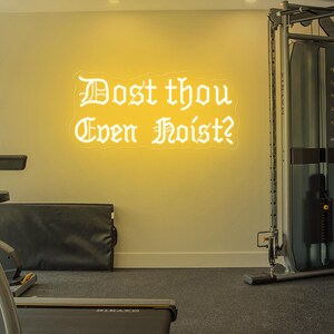 Puede incluir: Un letrero de ne&oacute;n amarillo con la frase "Dost thou Even Hoist?" en fuente g&oacute;tica. La imagen muestra equipos de gimnasio y un suelo negro.