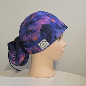 May include: A scrub cap with a vibrant purple and blue galaxy print. The cap features a ponytail design with a dark blue tie. A small white label with text is visible on the side. The cap is displayed on a mannequin head.
