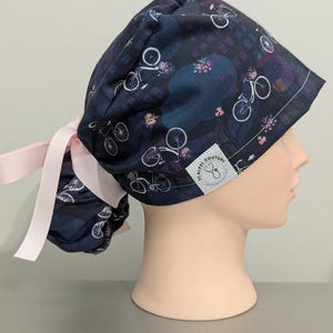 May include: A dark blue surgical scrub hat with a pink ribbon tie. The hat features a repeating pattern of bicycles and flowers. The hat has a white label with the text "SCALPEL COUTURE" and a small logo.