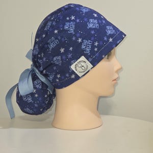 May include: A dark blue scrub cap featuring a repeating pattern of white flowers and the text "ORTHO SURGERY" in a white font. The cap includes a light blue ribbon tie at the back and a white rectangular label with the text "SCALPEL COUTURE".