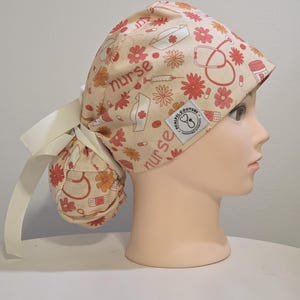 May include: A beige scrub cap with a floral and medical-themed pattern, including flowers, syringes, and the word "nurse." The cap has a white ribbon tie and a small label. The cap is on a mannequin head.