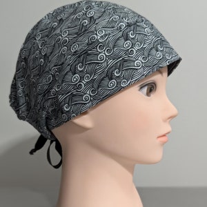 May include: A black and white patterned surgical scrub hat with a tie at the back. The pattern features a repeating design of waves and swirls.