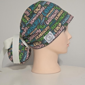 Puede incluir: Un gorro quirúrgico con un soporte para coleta, con un estampado de términos médicos como "Endoscopia" y "ERCP" en varios colores sobre un fondo gris. El gorro tiene una cinta blanca y una pequeña etiqueta con un símbolo médico.
