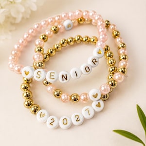 May include: Three stretch bracelets featuring a mix of light pink pearl-like beads, metallic gold beads, and white round beads with gold lettering. One bracelet spells out SENIOR with heart icons, and another displays the year 2027 with heart icons.