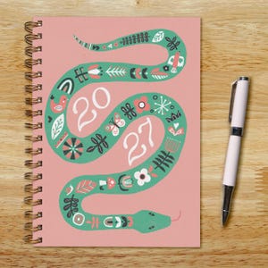 May include: A spiral-bound notebook with a pink cover featuring a large green snake illustration decorated with white and red floral and bird motifs. The numbers 20 and 27 are printed in white within the snake's curves. A white ballpoint pen is included.