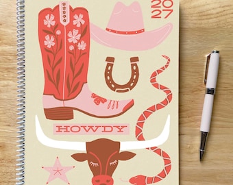 May include: A spiral-bound 2027 planner with a cream cover featuring a red and pink cowboy boot, a pink cowboy hat, a brown horseshoe, a red snake, a brown longhorn steer head, a pink star, and the text HOWDY in a red rectangular box.