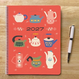 May include: A spiral-bound 2027 planner with a red cover featuring a pattern of various illustrated teapots and kettles in shades of blue, green, pink, and white, accented by small star shapes, accompanied by a white ballpoint pen with silver trim.