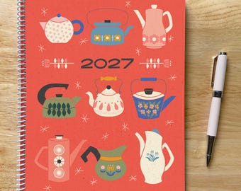 May include: A spiral-bound 2027 planner with a red cover featuring a pattern of various illustrated teapots and kettles in shades of blue, green, pink, and white, accented by small star shapes, accompanied by a white ballpoint pen with silver trim.