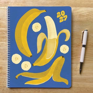 May include: A spiral-bound notebook with a blue cover featuring yellow banana illustrations and the text 2027, accompanied by a white pen. The notebook displays whole bananas, a peeled banana, and several sliced banana rounds in a pattern.