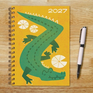 May include: A spiral-bound 2027 planner featuring a large green alligator illustration on a yellow cover, accented by white circular lily pad shapes. A white ballpoint pen with silver accents is positioned to the right of the notebook.