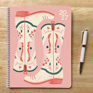 May include: A spiral-bound notebook with a pink cover featuring two cream-colored cowboy boots decorated with pink and dark green butterfly wing patterns and floral motifs. The text 2027 is printed in the upper right corner. A white ballpoint pen.
