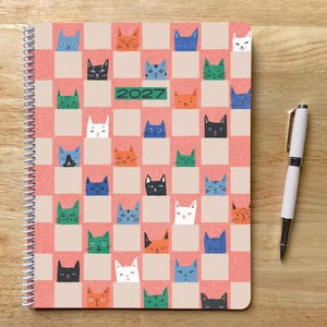 May include: A spiral-bound 2027 planner featuring a repeating checkerboard pattern of pink squares and beige squares, each containing a colorful illustrated cat face. A white ballpoint pen with silver accents is positioned to the right of the planner.