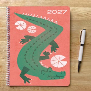 May include: A spiral-bound 2027 planner with a coral-colored cover featuring a large green crocodile illustration and white circular lily pad shapes. A white ballpoint pen with silver accents is positioned to the right of the planner.