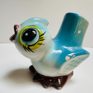 May include: A vintage ceramic bird figurine in shades of blue, white, yellow, and brown. The bird has large, expressive eyes with black pupils and yellow accents. The figurine has a glossy finish and is likely a decorative item.