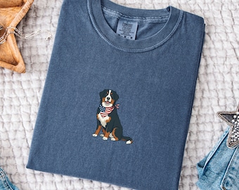 May include: A blue short-sleeved t-shirt featuring an embroidered graphic of a Bernese Mountain Dog wearing a red, white, and blue American flag patterned bandana. The shirt has a crew neckline and a label inside the collar reading Comfort Colors.