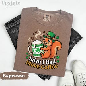 May include: A brown t-shirt with a graphic of a squirrel wearing a green hat, holding a coffee mug with shamrocks. The text "Irish I Had More Coffee" is printed below the graphic. The shirt is paired with dark denim jeans and white sneakers.