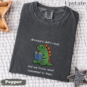 May include: A charcoal gray crew neck t-shirt featuring an embroidered green dinosaur wearing glasses and reading a blue book. The text above and below the dinosaur reads, "dinosaurs didn't read... and we know what happened to them."