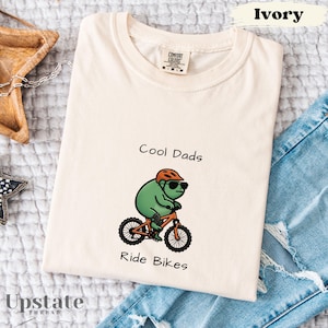 May include: Ivory-colored t-shirt with the text "Cool Dads Ride Bikes" and a cartoon frog riding a bicycle. The frog wears sunglasses and a helmet. The shirt is folded on top of blue jeans.