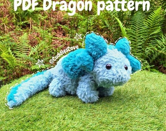 May include: A blue and turquoise plush dragon toy with a long tail. The toy is sitting on a green grassy surface. The text "PDF Dragon pattern" is written above the toy. The text "@CuddlesCove" is written below the toy.