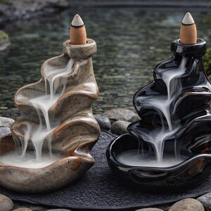 Könnte beinhalten: Zwei Räuchergefäße aus Keramik mit gestuftem Wasserfall-Design. Eines ist beige gesprenkelt, das andere glänzend schwarz. Beide haben einen Räucherkegel an der Spitze, von dem weißer Rauch kaskadenartig in das untere Becken hinabfließt.