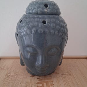 Pode incluir: Um queimador de óleo de cerâmica cinza em forma de cabeça de Buda. A peça possui uma tampa superior removível com uma pequena abertura circular e vários orifícios adicionais ao redor do topo da cabeça. A superfície tem um acabamento brilhante.