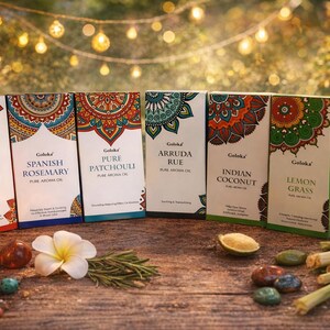 Pode incluir: Seis caixas de Goloka Pure Aroma Oil são exibidas, cada uma com um design de rótulo e nome de aroma exclusivos. Os aromas incluem Dragon Blood, Spanish Rosemary, Pure Patchouli, Arruda Rue, Indian Coconut e Lemon Grass. Uma vela acesa, flores e cristais também estão presentes.