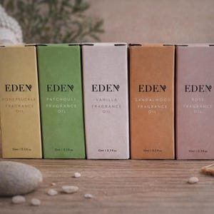 Pode incluir: Cinco caixas de óleos de fragrância em várias cores, cada uma rotulada com o nome de uma fragrância e a marca "EDEN". As caixas estão dispostas sobre uma superfície de madeira, com uma estátua de Buda e uma vela ao fundo. Cada caixa contém 10 ml de óleo de fragrância.