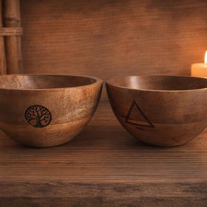 Peut inclure: Deux bols en bois avec une finition naturelle sont présentés sur une surface en bois. L'un des bols présente un motif d'arbre de vie, tandis que l'autre porte un symbole triangulaire. Des bougies allumées et une pile de pierres sont en arrière-plan.