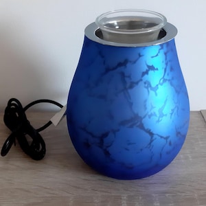 Peut inclure: Diffuseur d'huiles essentielles bleu avec un motif marbré. Il possède un bol en verre transparent et un bord argenté. Un cordon d'alimentation noir est connecté. Il est posé sur une surface en bois.