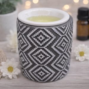 Può includere: Un diffusore di oli essenziali con motivo grigio e bianco con una ciotola in ceramica bianca. Il diffusore è cilindrico e presenta un design geometrico. Due piccole bottiglie di olio essenziale marrone sono sullo sfondo, insieme a fiori bianchi.