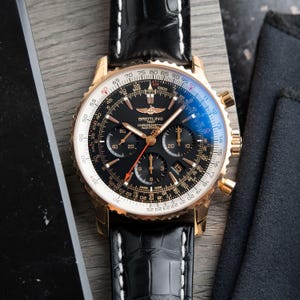 May include: A Breitling Navitimer chronograph watch with a rose gold case, black dial, and black leather strap with white stitching. The dial features three sub-dials, a date window, and a slide rule bezel with white and black markings.