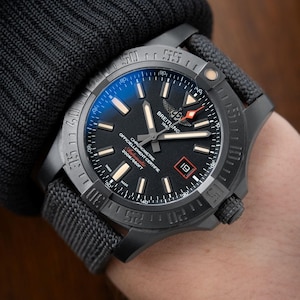 May include: A black Breitling wristwatch with a matte finish, featuring a dark dial, luminous hour markers, a date window at the three o'clock position, and a textured gray fabric strap. The watch face displays the Breitling logo and text.