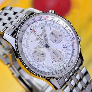 May include: A silver-tone Breitling Navitimer chronograph watch with a white dial, three sub-dials, a date window at the three o'clock position, and a metallic link bracelet. The watch face features a slide rule bezel with black markings.