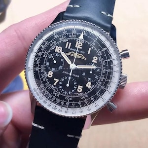 Könnte beinhalten: Eine Breitling Navitimer Chronographenuhr mit schwarzem Zifferblatt, weißer Rechenschieber-Lünette und leuchtenden Zeigern. Die Uhr verfügt über drei Hilfszifferblätter, arabische Stundenziffern und ein schwarzes Lederarmband mit weißen Kontrastnähten.