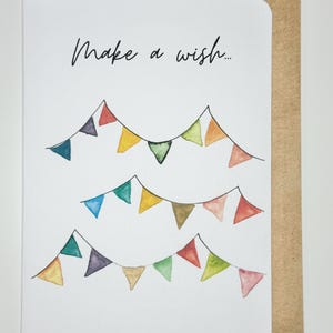 Könnte beinhalten: Weiße Grußkarte mit dem Text "Make a wish..." in schwarzer Schrift. Die Karte zeigt drei Reihen bunter Aquarell-Wimpelbanner. Ein brauner Umschlag befindet sich hinter der Karte.