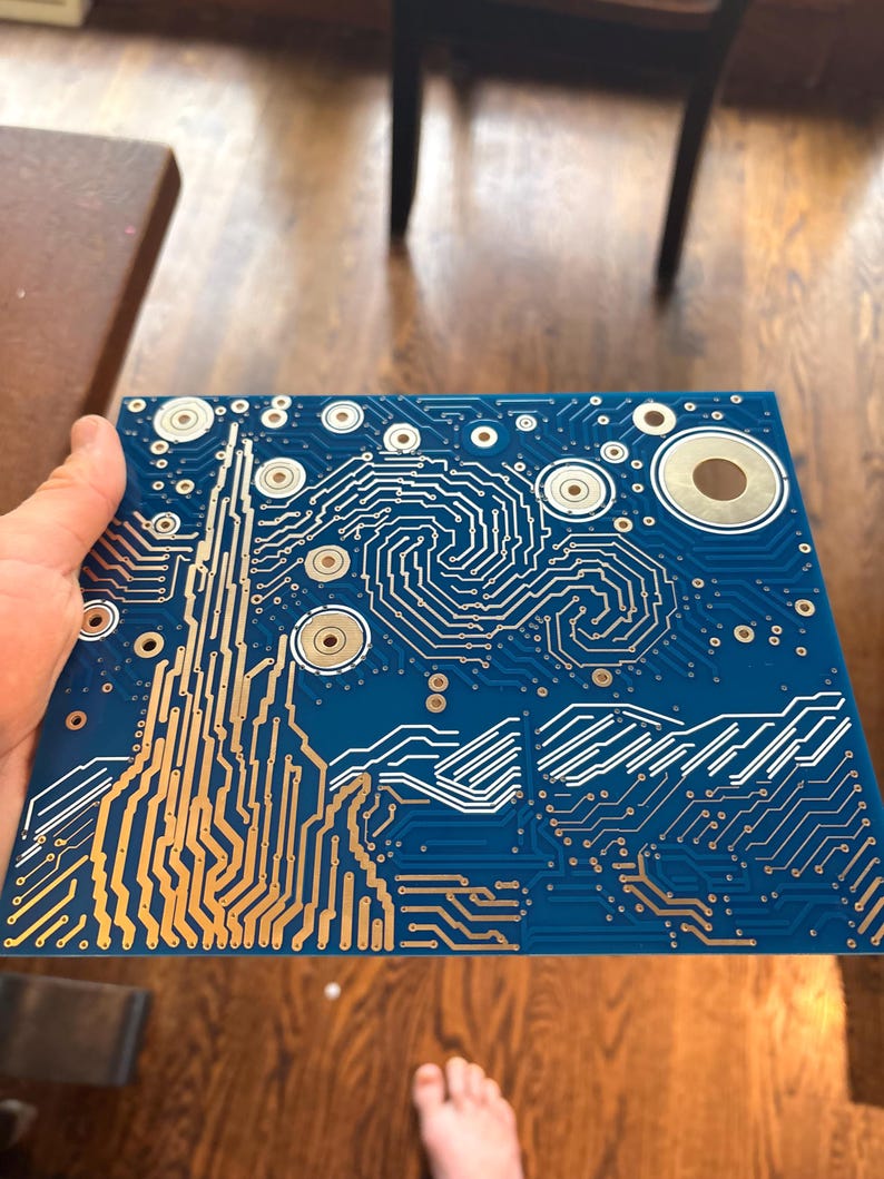 Puede incluir: Una placa de circuito impreso cuadrada de color azul con un patr&oacute;n de pistas conductoras de color cobre que replica el cielo arremolinado y el paisaje de una pintura famosa, con puntos de contacto met&aacute;licos circulares.