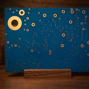 Puede incluir: Un panel rectangular azul con un patr&oacute;n disperso de anillos circulares dorados de varios tama&ntilde;os, apoyado sobre una base de bloque de madera horizontal. El dise&ntilde;o recuerda a un cielo nocturno estrellado.