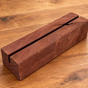 Puede incluir: Un bloque de madera rectangular de tono marr&oacute;n rojizo intenso con una veta visible. El bloque presenta una ranura vertical larga y estrecha a lo largo de la superficie superior, que tambi&eacute;n muestra una peque&ntilde;a grieta natural cerca del centro.