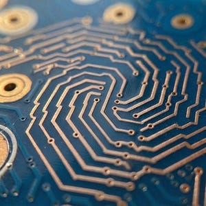 Puede incluir: Una vista de cerca de una placa de circuito impreso azul con intrincadas rutas de cobre dispuestas en un patr&oacute;n en espiral. La placa incluye varias almohadillas de contacto de cobre circulares con orificios centrales, mostrando el dise&ntilde;o electr&oacute;nico detallado.