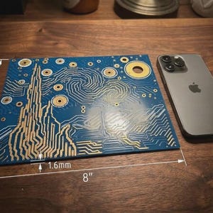 Puede incluir: Una placa de circuito impreso azul con un patr&oacute;n art&iacute;stico dorado de l&iacute;neas y c&iacute;rculos en espiral, que mide 16,51 cm por 20,32 cm con un grosor de 1,6 mm, junto a un tel&eacute;fono inteligente gris sobre una superficie de madera.