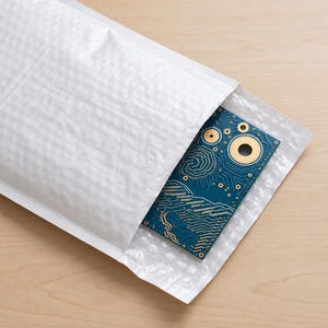 Puede incluir: Una placa de circuito impreso rectangular azul con puntos de contacto circulares dorados y caminos de cobre intrincados, parcialmente insertada en un sobre de env&iacute;o blanco con burbujas de polietileno.