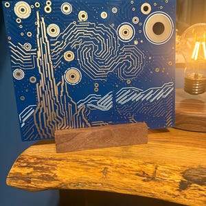 Puede incluir: Una placa de circuito rectangular azul con un patr&oacute;n de trazas de color dorado que imita la composici&oacute;n arremolinada de un cielo estrellado. La placa est&aacute; montada verticalmente en un peque&ntilde;o soporte de bloque de madera rectangular, mostrando intrincadas l&iacute;neas met&aacute;licas.