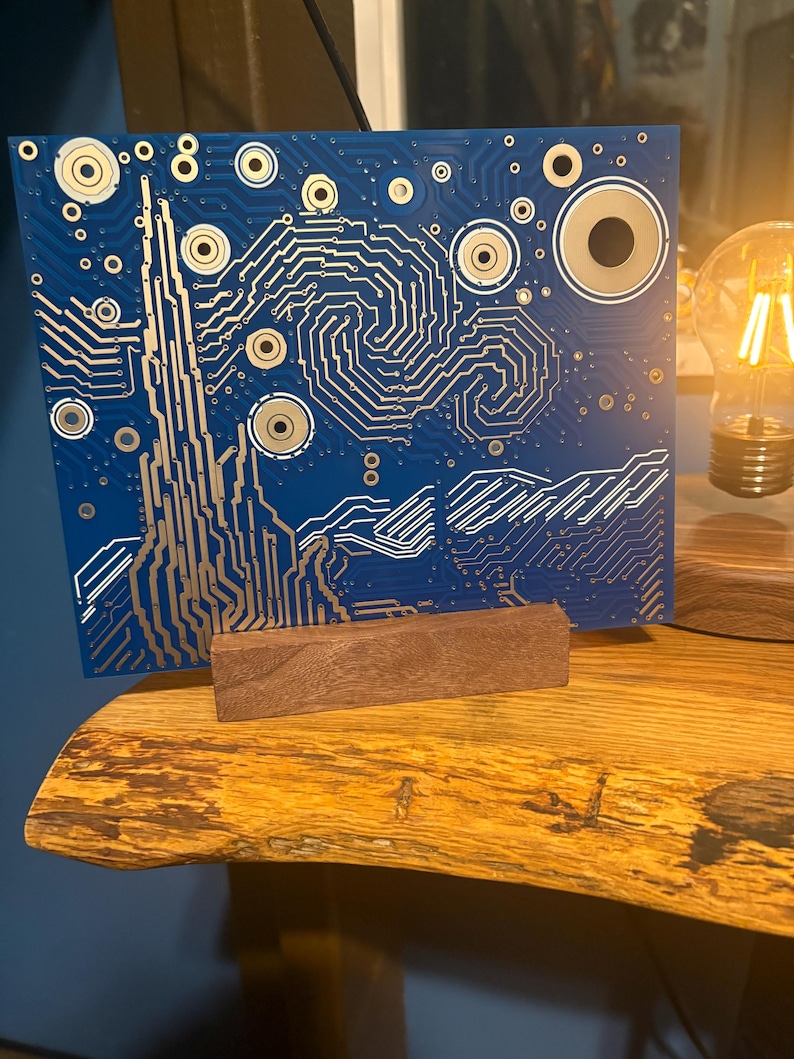Puede incluir: Una placa de circuito rectangular azul con un patr&oacute;n de trazas de color dorado que imita la composici&oacute;n arremolinada de un cielo estrellado. La placa est&aacute; montada verticalmente en un peque&ntilde;o soporte de bloque de madera rectangular, mostrando intrincadas l&iacute;neas met&aacute;licas.