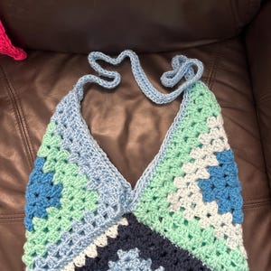 May include: A crocheted halter top with a geometric pattern in shades of blue, green, and white. The top is made with granny squares and has a v-neckline.