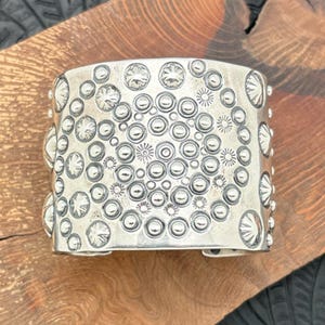 May include: A wide, silver cuff bracelet with a detailed pattern of raised circles and starburst designs. The bracelet has a slightly curved shape and is made of a shiny, metallic material. The cuff is displayed on a wooden surface.