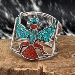 May include: Silver cuff bracelet with a turquoise and red inlay design. The bracelet features a stylized figure with outstretched wings, crafted with small turquoise stones and red inlay. The openwork design is set against a wooden surface.