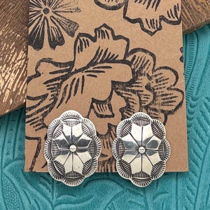 May include: Pair of silver-toned stud earrings. Each earring features a floral design with a central starburst pattern and a scalloped edge. The earrings are displayed on a brown card with a floral print.