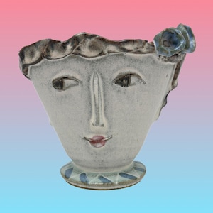 Puede incluir: Un jarrón de cerámica con forma de rostro en gris claro. El jarrón presenta un rostro estilizado con ojos oscuros, una nariz larga y labios rojos. Una flor azul adorna el lateral, y el borde tiene un borde texturizado gris oscuro.