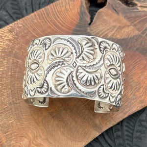 May include: A wide, silver cuff bracelet with an intricate, embossed floral design. The cuff features a series of stylized flower and leaf patterns, creating a textured surface. The bracelet is displayed on a wooden surface.